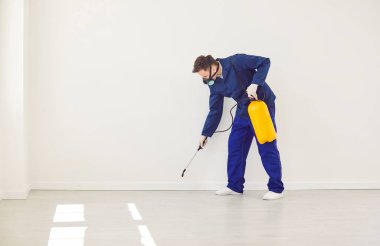 Genç bir erkek müteahhit boş bir odada koruyucu maske takıp dezenfektan spreyle zehir püskürtüyor. Dezenfeksiyon ve koruma kavramı.