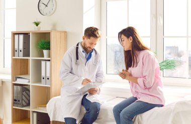 Klinikte ya da hastanede kadın hastayla konuşan bir erkek doktor. Beyaz önlüklü, steteskoplu bir erkek doktor muayene odasında genç bir kadınla oturuyor, tedaviden bahsediyor, panoya yazı yazıyor.