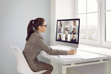 Uzak çalışanlarından oluşan bir ekiple çalışan ciddi bir iş kadını. Takım elbiseli genç bir kadın ofis yerinde modern masaüstü bilgisayarıyla masada oturuyor, online iş toplantısı yapıyor ve notlar alıyor.