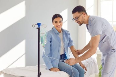 Genç, dost canlısı erkek hemşire, hastanede kadınlara serum ya da vitamin terapisi almalarında yardım ediyor. Klinikteki kanepede oturan kadın enjeksiyon terapisi görüyor. Sağlık hizmeti kavramı.
