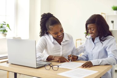 Afro-amerikan ofis dostu işçiler, yeni projede işbirliği yapan kadınlar, laptopla çalışma masasında oturmak, gülümsemek, yeni öğrencilere öğretmenlik yapmak, belgeye işaret etmek, profesyonel akıl hocalığı