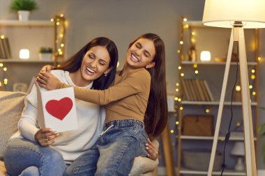 Aile içinde ebeveyn ve çocuk ilişkisi. Neşeli genç kız, annesine anneler günü kartını sevgiyle sunar. Mutlu kadın kızının selamlarını okuyor ve neşeyle gülüyor..