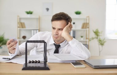 Grumpy bored overworked businessman looking unhappily at stack of paper work, sitting at work desk with Newtons cradle pendulum at foreground, having no motivation. Burnout and overwork concept. clipart