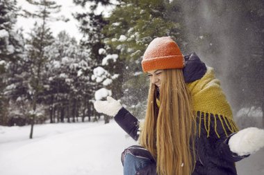 Neşeli mutlu genç kadın ormanda ya da parkta eğleniyor ve soğuk kış gününün tadını çıkarıyor. Karlı ormanda eğlenirken ağaçlardan kar yağarken oturup kahkahalarla gülen kızın portresi..