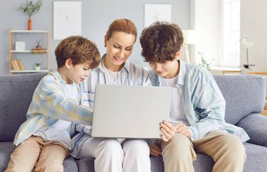 Kanepede oturan iki çocuklu mutlu genç anne birlikte dizüstü bilgisayar kullanıyor. Çocuklarla kanepede dinlenen gülümseyen kadın hafta sonunun keyfini çıkarıyor. İnternetten komik bir çizgi film izliyor ya da görüntülü konuşuyor..