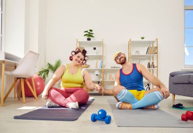 Komik şişman kadın ve sakallı adam spor kıyafetleri içinde el ele tutuşup spor sonrası egzersiz sonrası kameraya bakıyor. Evde yoga minderinde oturuyorlar..