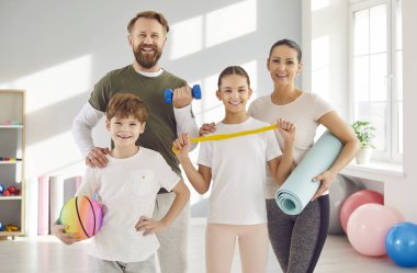 Dört kişilik mutlu bir aile birlikte spor yapıyor. Neşeli, kendinden emin anne, baba ve çocuklar spor salonunda ellerinde spor malzemeleriyle kameraya bakıyor ve gülümsüyorlar.