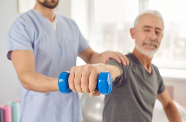 Jimnastik salonundaki profesyonel fizyoterapist yaşlı adama kollar, omuzlar ve dirsekler üzerine egzersizler yaptırıyor. Fizyoterapi rehabilitasyon konsepti. Dambılı tutarken nazikçe odaklan.