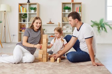 Mutlu ebeveynler kızlarıyla vakit geçirip tahta bloklarla eğitici oyunlar oynuyorlar. Anne, baba ve anaokulu çocuğu birlikte kule inşa etmek için taş diziyorlar. Evde aile aktiviteleri.