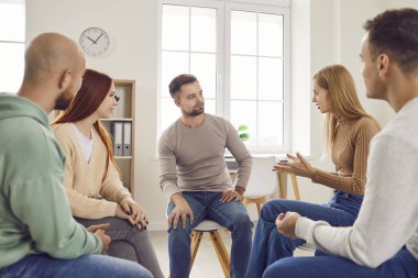 Çeşitli gençlerden oluşan bir grup, bir ekip toplantısı sırasında bir çemberin içinde oturup tartışmalara dalıyorlar. Takım çalışmasının ve iletişimin sinerjisi, takım halinde konuştukları ve fikir alışverişinde bulundukları sürece açıktır..