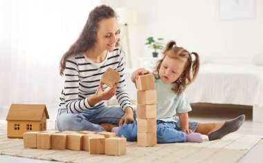 Anne ve çocuk, evde birlikte ahşap küplerle bir kule inşa ediyorlar, boş zamanların keyfini çıkarıyorlar. Paylaşılan oyun zamanı yakınlığı teşvik eder, ev ortamında neşeli anlar yaratır..