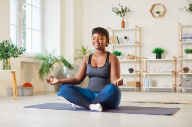 Sportif yogiler orta yaşlı mutlu Afrikalı Amerikalı kadın. Nilüfer çiçeğinde huzur içinde oturuyor. Jest parmaklarıyla poz veriyor, derin düşünceli meditasyonun tadını çıkarıyor.