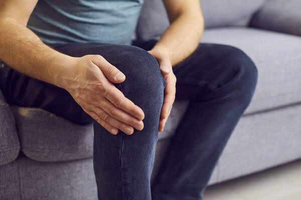 Close up man holding, massaging, touching knee with hands, feeling pain, suffering from ache in leg, sitting on couch at home, feeling unwell, health problem concept, osteoarthritis, health problem 
