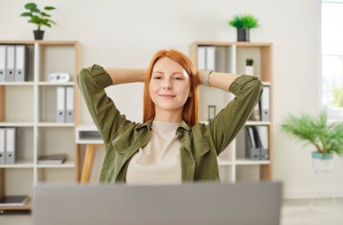 Laptopta tasasız genç, kızıl saçlı bir kız, yoğun bir iş gününden sonra dinlenen bir çalışan, yaslanmış bir ofis koltuğu, elleri başının arkasında, yapılan işten memnun olmak için gülümseyen, başarılar, iş sonuçları.
