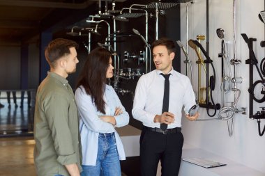 Dost canlısı gülen bir satıcı müşterilerle konuşuyor, farklılıkları anlatıyor ve banyoları için duş başlığını seçmelerine yardım ediyor. İnşaat pazarında satış asistanının yanında duran genç çift..