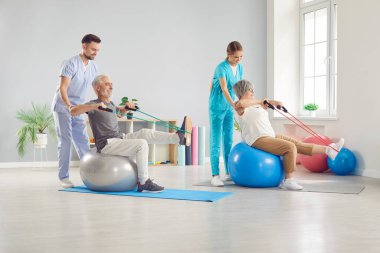 Profesyonel fizyoterapistler spor malzemeleriyle spor yaparken kıdemli hastalara yardım ederler. Mutlu yaşlı çift, fizyoterapi kliniğinde elastik bantlarla İsviçre fitbol topları üzerinde egzersiz yapıyor.