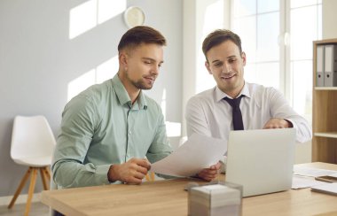 İki erkek meslektaşı iş yerinde parlak ofiste belgeleri tartışıyorlar. Genç Kafkas erkek ofis çalışanları ofis odasında dizüstü bilgisayarla masada otururken belgeleri inceliyorlar..