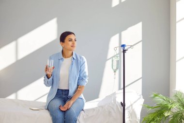 Young smiling woman sitting on the couch in medical clinic looking at the window with a glass of water while receiving IV drip infusion and vitamin injection therapy. Healthcare concept. clipart