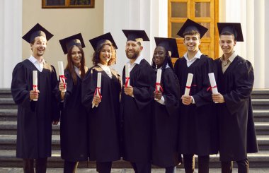 Bir grup mutlu, çok ırklı uluslararası mezun öğrencinin portresi üniversite mezuniyet elbisesi giymiş ve açık havada diploma tutuyorlar. Eğitim ve mezuniyet kavramı.