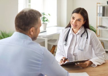 Tıbbi muayenehanedeki hasta erkek portresi masasında oturan ve elinde randevu ile rapor tutan ve klinikteki tıbbi muayene sırasında danışmanlık yapan dost canlısı bir kadın doktoru dinliyor..