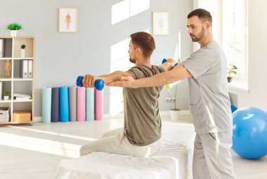 Rehabilitasyon kliniğinde kanepede oturan hasta hemşirelerle halterli spor egzersizleri yapıyor. Fizyoterapist hastaya dambıl kaldırmada yardım ediyor. Fiziksel rehabilitasyon kavramı.