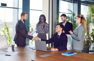Patron, ofiste başarılı bir anlaşmayı kutlayan meslektaşlarıyla birlikte toplantı sırasında bir çalışanın elini sıkıyor. Anlaşma, işbirliği ve ekip içindeki topluluk..