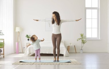 Anne ve çocuğun boş vakitlerinde yoga çalışmaları var. Mutlu güzel genç kadın ve sevimli küçük kızı evdeki beyaz yatak odasının iç kısmında egzersiz minderinde yan yana duruyorlar. Aile kavramı