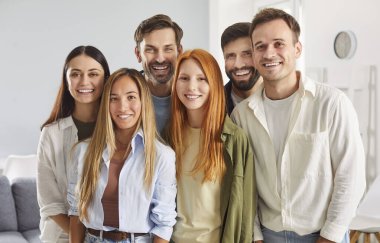 Mutlu gülümseyen bir grup genç arkadaş ya da sıradan kıyafetler giymiş iş arkadaşları yan yana duruyor ve içeride kameraya neşeli ve pozitif bakıyorlar. Dost canlısı erkek ve kadınların portresi.