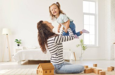 Neşeli anne, ahşap küplerle küçük ahşap evin arasında oturmuş anaokulu kızını kucağında tutuyor ve onu uçuruyor. Mutlu aile anne ve çocuğu, geniş yatak odasında birlikte eğleniyorlar..