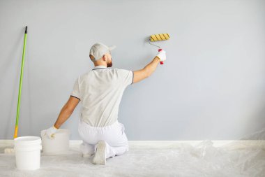 Profesyonel bir tamircinin arkası, bir ev yenileme projesi sırasında duvar boyamak için paten fırçası kullanır. Adam inşaat işinde uzmanlık gösteriyor, iş sorumluluklarını yerine getiriyor..