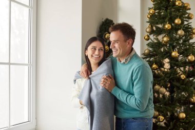 Mutlu gülümseyen genç bir adamın portresi kız arkadaşını sıcak bir battaniyeyle örterek Noel ağacının yanında duruyor. Aşık mutlu bir çift. Kış tatili ve yeni yıl konsepti.