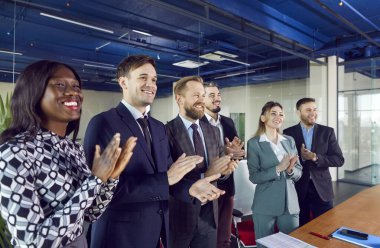 Bir grup mutlu, çok ırklı iş adamı konferans odasında durmuş başarılı grup toplantısından sonra konuşmacıyı alkışlıyorlar. Mesleki eğitim, iş toplantısı, sunum veya koçluk.