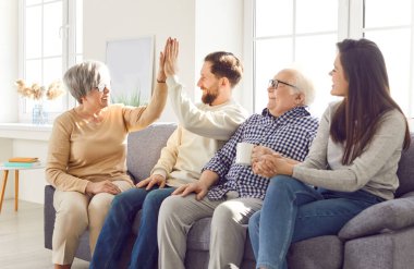 Mutlu gülümseyen yaşlı ebeveynler ve onların yetişkin çocukları evdeki oturma odasında çay içip beşlik çakarak birlikte vakit geçiriyorlar. Aile eğlence konsepti.
