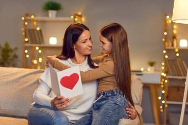 Neşeli anne, tatlı kızıyla birlikte evdeki kanepede oturuyor ve tatilde anneler gününde kalpten el yapımı tebrik kartı okuyor. Mutlu kadın kızından hediye alıyor..