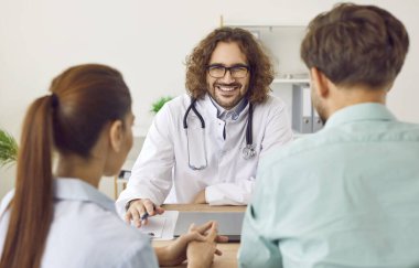 Dost canlısı erkek doktor, evli çifte hamilelik yönetimi tavsiyesinde bulunuyor ve onlara doğum öncesi tavsiyelerde bulunuyor. Gülümseyen erkek doğum uzmanı jinekolog danışman çiftin portresi..