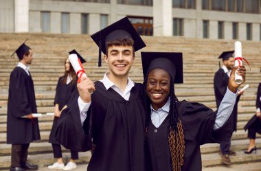 Üniversite mezuniyet elbisesi giymiş, elinde diplomasıyla gülümseyen genç bir adam ve kadının portresi. Sınıf arkadaşlarının arka planında duran kendinden emin mutlu öğrenciler. Mezuniyet ve eğitim kavramı.