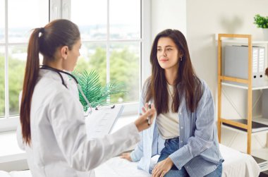 Güzel genç bayan doktoru dinliyor, yakın plan. Doktor bir dosya tutuyor ve bir kadınla konuşuyor. Klinikte danışmanlık ve teşhisler, ön sağlık kontrolü