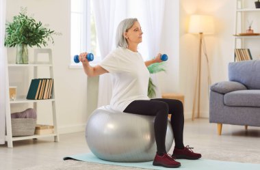 Yaşlı kadın evde spor salonuna gider, dambıl kullanır ve jimnastik için spor topu kullanır. Spor, pilates ve aktif kalmaya odaklanmış, tanıdık bir alanda egzersiz yapmaktan hoşlanıyor..