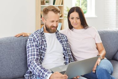 Oturma odasındaki kanepede birlikte oturan genç bir çiftin portresi sosyal medya haberleri veya komik videolar için dizüstü bilgisayar kullanıyor. Gülümseyen adam ve kadın kanepede dinleniyor hafta sonunun tadını çıkarıyor.