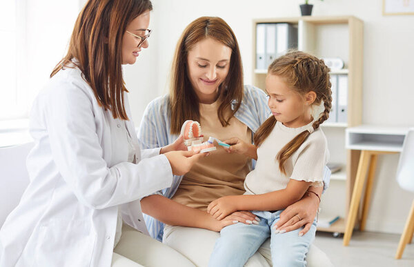 Dentist doctor or nurse teaches a mother and daughter proper oral hygiene in a hospital setting. The healthcare professional demonstrates how to clean teeth with a toothbrush, promoting dental health.