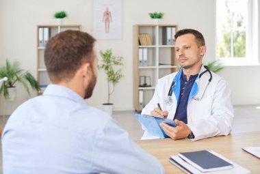 Laboratuvar önlüklü ciddi odaklı erkek doktor elinde dosya ile masada oturuyor ve hastaların şikayetlerini dikkatlice dinliyor. Klinikte sağlık kontrolü sırasında doktorla ya da kardiyologla konuşan adam.