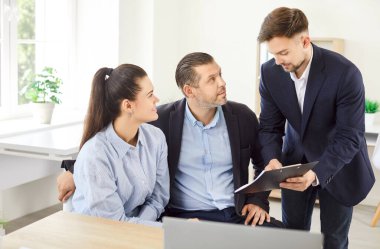 Profesyonel emlakçı müşterilere sözleşmenin şartlarını açıklıyor. Aile bireyleri ofis masasında oturuyor, dikkatle dinliyor, ev alıyor ya da kiralıyor, emlak acentesinde sözleşme imzalıyor..