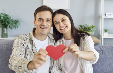 Neşeli, gülümseyen esmer kadın ve elinde kağıt kalp tutan adam evdeki oturma odasında kanepede oturuyorlar. Aşık mutlu bir çift. Romantik, ilişki ve Sevgililer Günü konsepti.