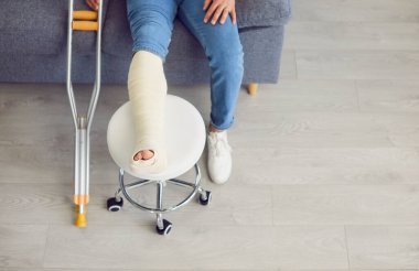 Koltuk değneklerinin ve alçıdaki kırık bacağın görüntüsü. Ayağı ve bacağı yaralı genç bir adam evdeki kanepede oturuyor, ayağı taburede dinleniyor. Kesilmiş çekim, üst görünüm.