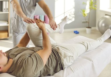 Erkek doktor ya da osteopat masajı ve klinikte rehabilitasyon sırasında hasta bacağını esnetme. Fizyoterapist, kanepede genç bir adama diz tedavisi uyguluyor..