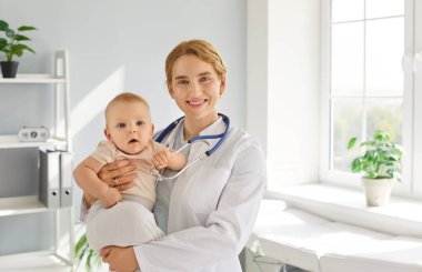 Klinikte ya da hastanede tıbbi muayene sırasında küçük bebek hastayı elinde tutan gülümseyen bir kadın doktor portresi. Pediyatrik ve sağlık hizmetleri kavramı.