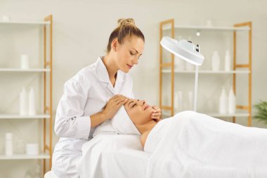 Profesyonel kadın güzellik uzmanı ya da kozmetik uzmanı güzellik salonunda özel lambası olan genç ve güzel bir kızın cilt durumunu inceliyor. Yüz masajı, cilt bakımı ve tedavi konsepti.