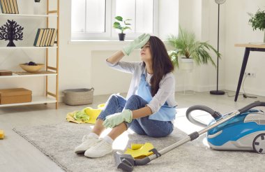 Evde temizlik yaptıktan sonra yerde oturan yorgun ev hanımı. Lastik eldivenler giymiş üzgün genç bir kadın, bitmek bilmeyen ev işlerinden bıkmış, oturma odasındaki halıya oturmuş, alnındaki teri siliyor.