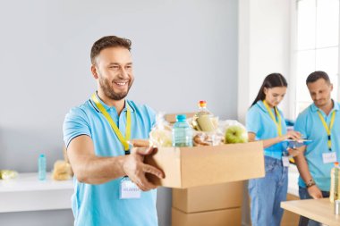 Neşeli arkadaş canlısı erkek gönüllü, insani yardım merkezinde ihtiyacı olan mültecilere yiyecek kutusunu veriyor. Ekibi de arka plan üzerinde çalışıyor. Yardım organizasyonu ihtiyacı olanlara yardım sağlıyor..