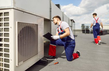 Beyaz erkek işçi çatıda bir havalandırma sisteminin denetimini, denetimini ve bakımını yapıyor. Havalandırma onarımı ve servis görevleri, verimli havalandırma hizmeti.